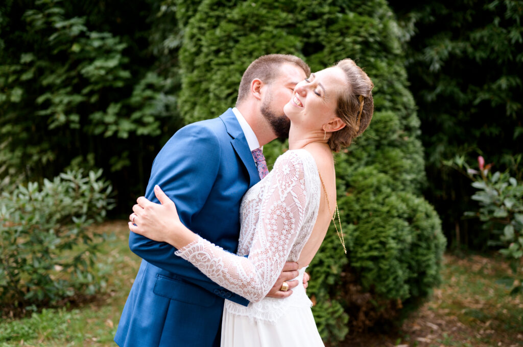 photo de couple souriant et heureux, mariage à Toulouse, Haute Garonne. Le marié fait un bisou dans le cou de la mariée. La mariée le tient dans ses bras et sourit. Elle porte une magnifique robe avec un collier de dos sur 2 rangs, robe signée carrière Mariage à Villefranche de Lauragais. Le marié fait un bisou dans le coup à la marié. Elle fait un grand sourire. Elle a une magnifique robe ornée d'un collier de dos de 2 rangs, Robe Carrière Mariage à Villefranche de Lauragais