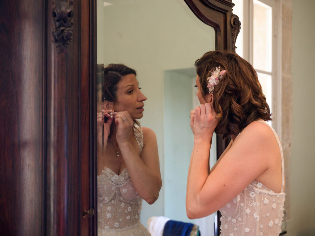 Préparatifs mariage Carcassonne la future mariée a changé de tenue. elle porte une robe pailletée Pronovia, elle se regarde dans un miroir, dans une chambre du Domaine de Russol. Elle met ses boucles d'oreilles avant la cérémonie laïque. Elle porte une broche en fleurs séchées dans les cheveux, par Rose & Marguerite à Montolieu.